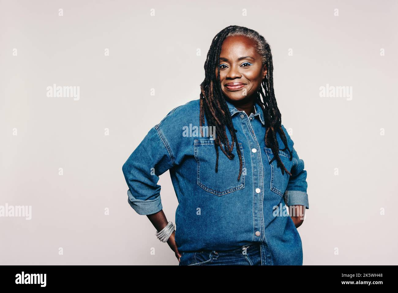 Donna matura alla moda che guarda la fotocamera mentre si trova su sfondo grigio. Donna elegante con lucchetti che indossa una giacca in denim e make-u Foto Stock