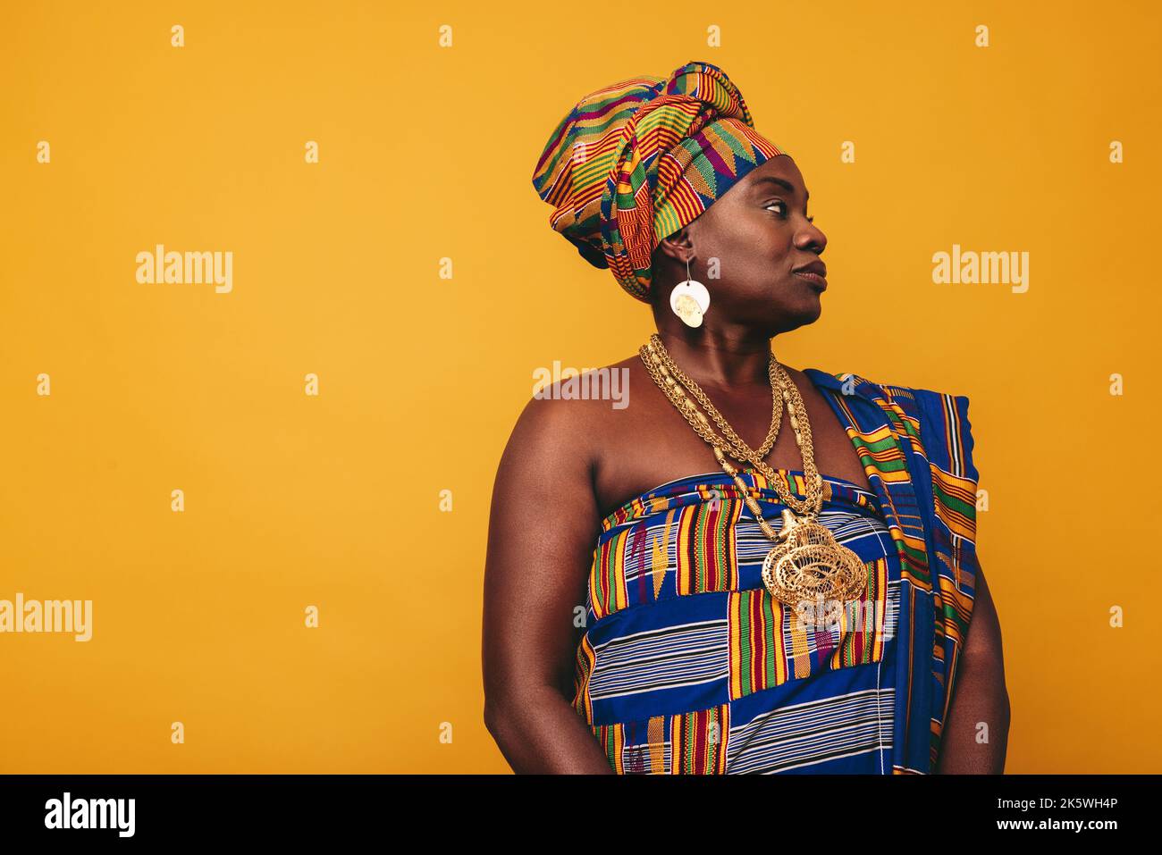 Elegante donna africana che indossa un abito tradizionale su sfondo giallo. Donna matura nera vestita di tessuto colorato Kente e gioielli d'oro Foto Stock