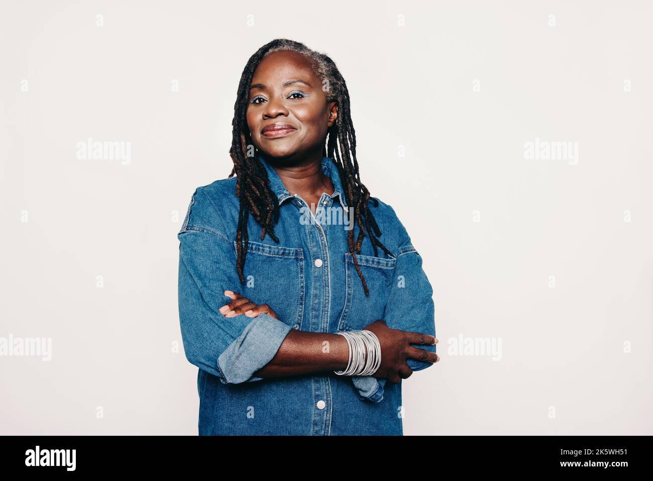 Donna matura ed elegante che guarda la fotocamera mentre si trova su uno sfondo grigio con le braccia incrociate. Donna alla moda con Dreadlocks che indossa un Foto Stock