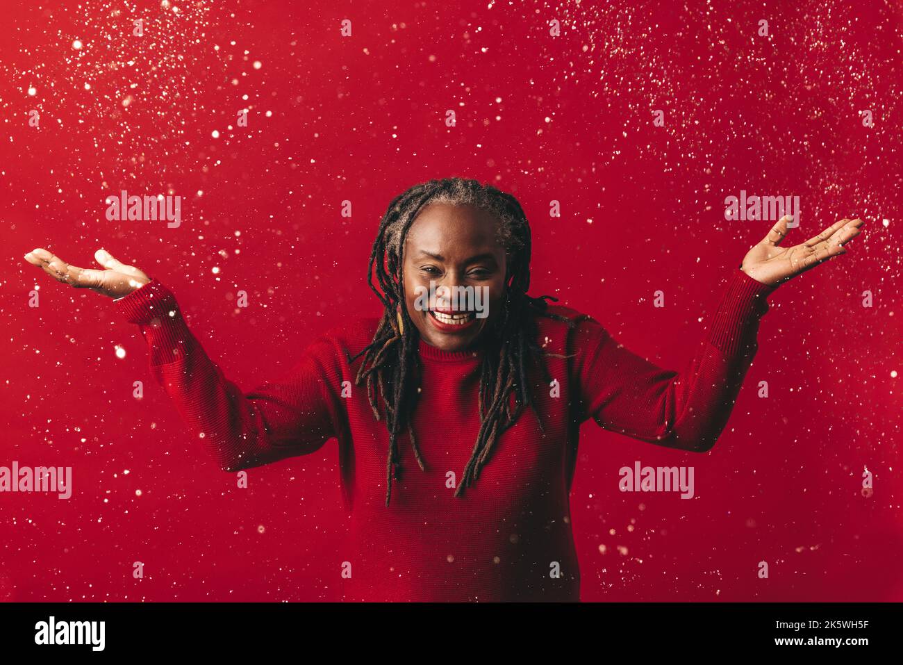 Donna eccitata con dreadlocks sorridente alla macchina fotografica mentre sta in piedi sotto i coriandoli cadenti. Donna matura felice che celebra la vita mentre si è in piedi contro Foto Stock