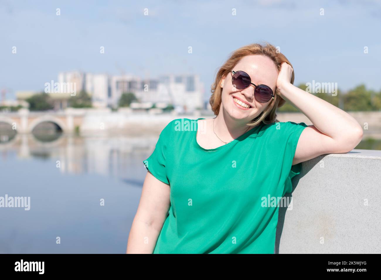Ritratto di una ragazza in occhiali da sole, una blusa verde in piedi sulla riva del fiume tenendo i capelli con la mano, copia spazio. Una ragazza sicura illuminata da Foto Stock
