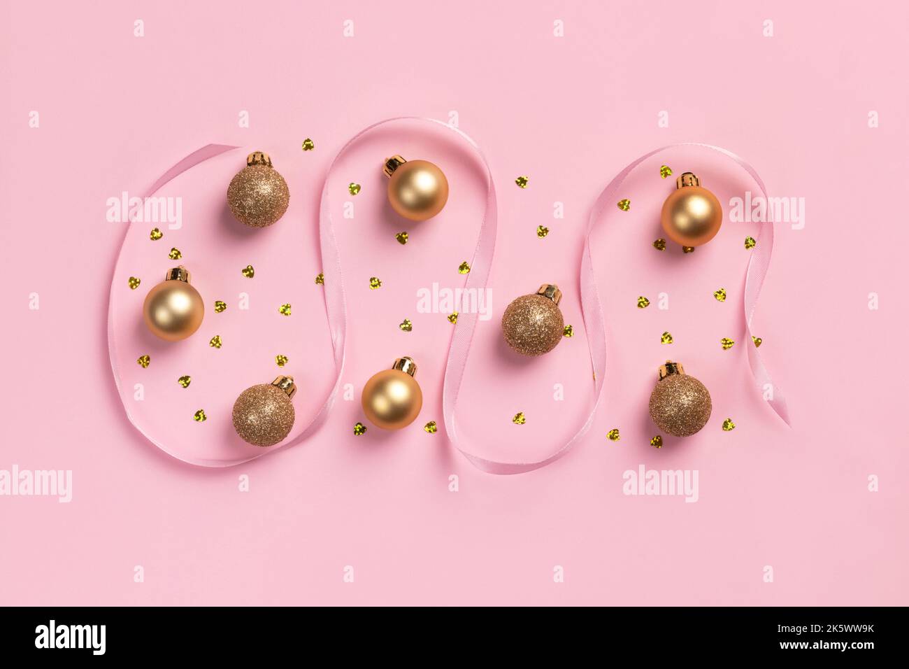 Cartolina di Natale con palline dorate, coriandoli a forma di cuore e nastro di raso su sfondo rosa Foto Stock