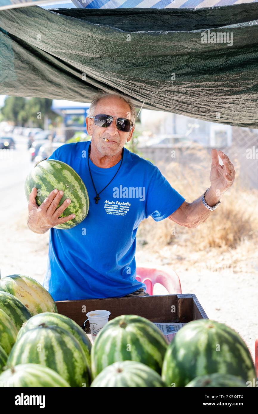 Una bancarella a bordo strada che vende cocomeri freschi coltivati localmente, Citrullus lanatus. Il felice supporto jovial stalla posa mentre tiene un grande anguria Foto Stock