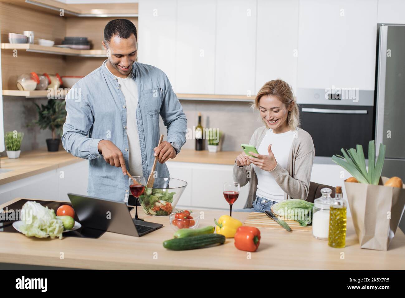 Ritratto di coppia multinazionale amorevole cucinare insalata insieme su una cucina. Allegra donna bionda che usa il telefono cellulare per trovare la ricetta di insalata vegana mentre suo marito mescola le verdure in una ciotola di vetro Foto Stock