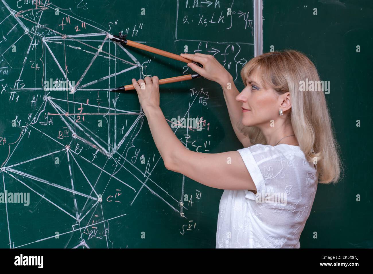 Una giovane maestra di matematica caucasica si trova nella lavagna e spiega la soluzione di un problema matematico Foto Stock