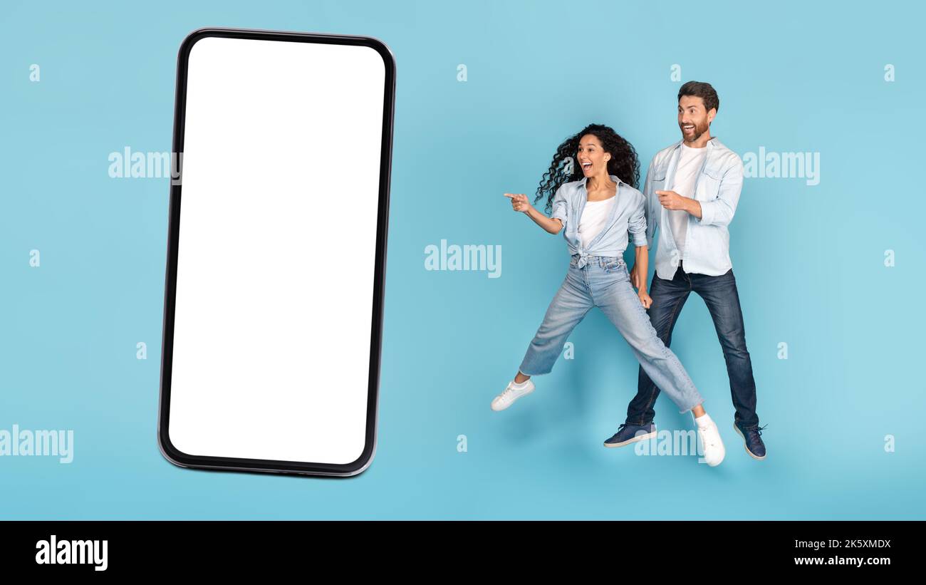 Scioccato giovane uomo caucasico e ragazza araba saltano in aria, punta il dito al telefono con spazio vuoto Foto Stock