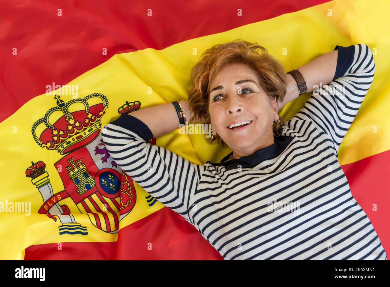 Donna anziana sorridente sdraiata sulla bandiera rossa e gialla della Spagna. Orgoglio spagnolo. Giorno della hispanidad. Columbus giorno Foto Stock