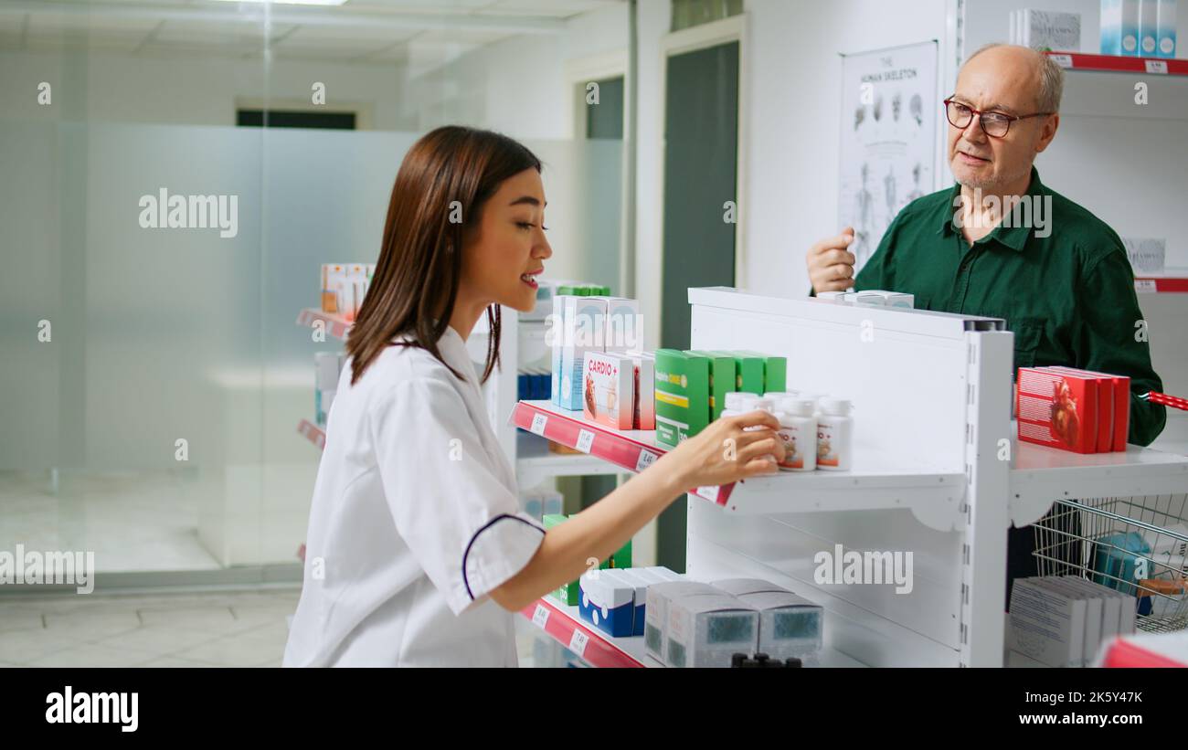 Farmacista asiatico aiutare diversi clienti con la malattia, dando prodotti farmaceutici da scaffali farmacia. Dipendente che fornisce assistenza medica alle persone, spiegando farmaci in farmacia. Foto Stock