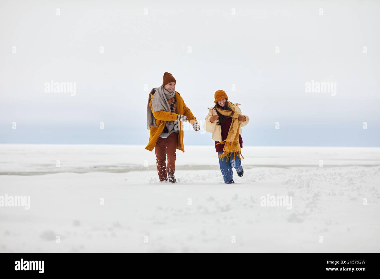 Vista frontale ritratto di giovane coppia spensierata che corre verso la fotocamera all'aperto in inverno da lago ghiacciato, copia spazio Foto Stock
