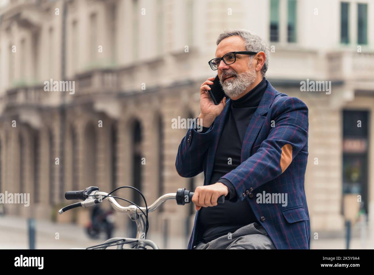 Imprenditore maschio caucasico anziano che trascorre attivamente il suo tempo libero. In bicicletta attraverso il centro della città. Uomo bearded grigio-capelli in occhiali con cornice nera, e blazer controllato parlando tramite smartphone. Foto di alta qualità Foto Stock