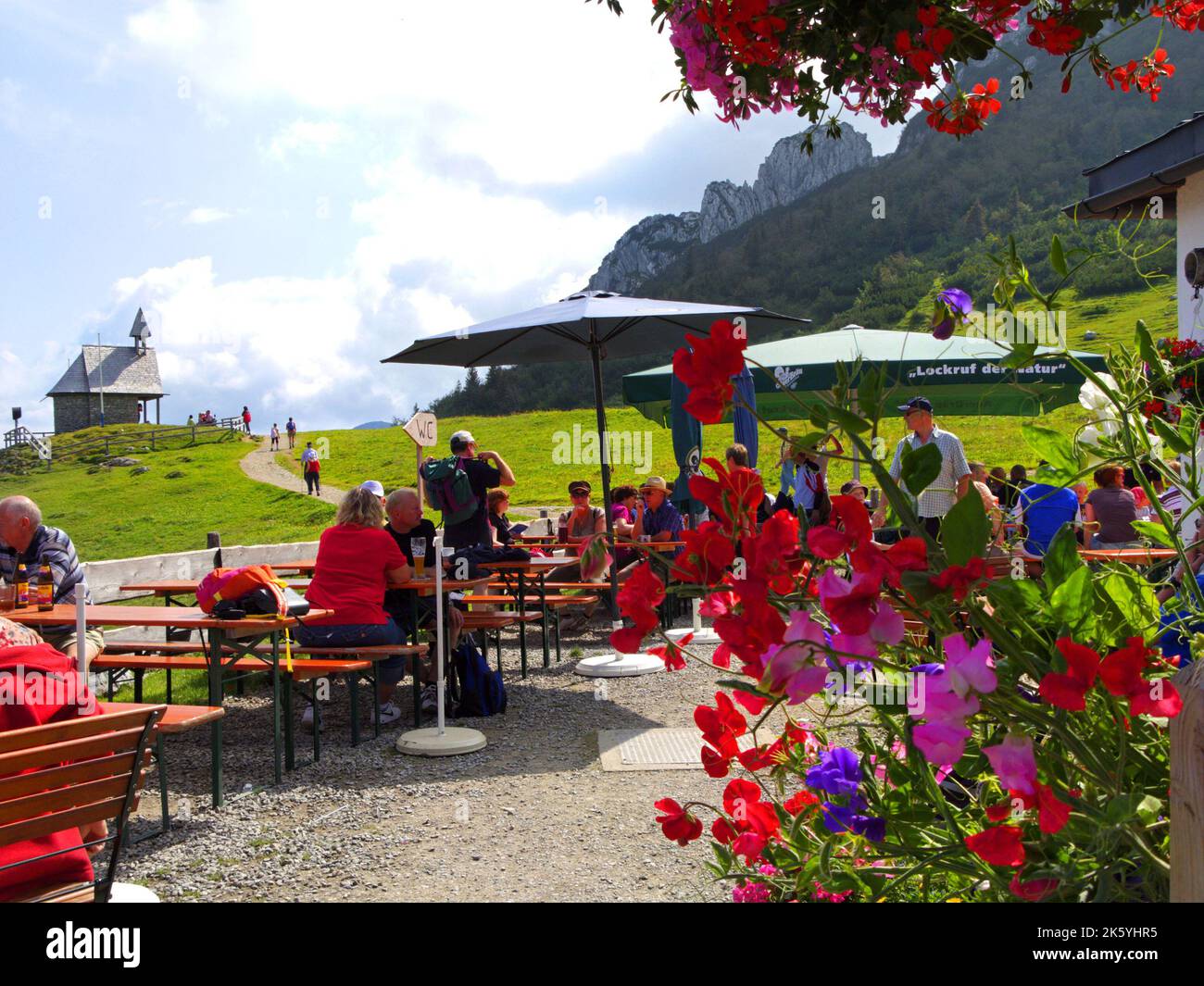 Terrazza di un ristorante alpino tedesco 'Steinling Alm' sulla montagna Kampenwand, ospiti, fiori e cappella, Chiemgau, Baviera, Germania. Terrasse des Alp Foto Stock