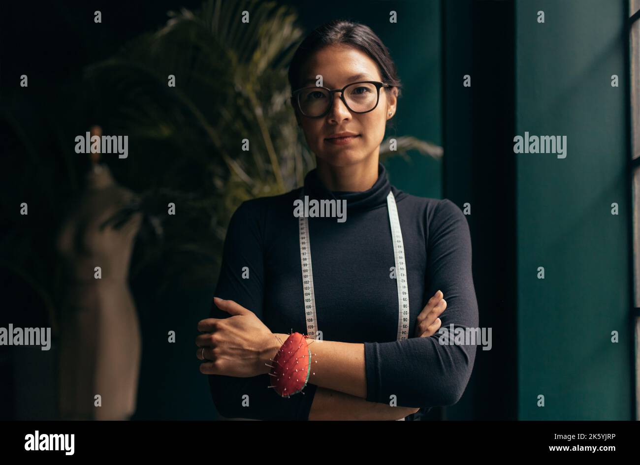 Ritratto di una stilista di moda femminile che guarda la fotocamera mentre si trova sul posto di lavoro. Donna in piedi in studio di moda con le braccia incrociate. Foto Stock
