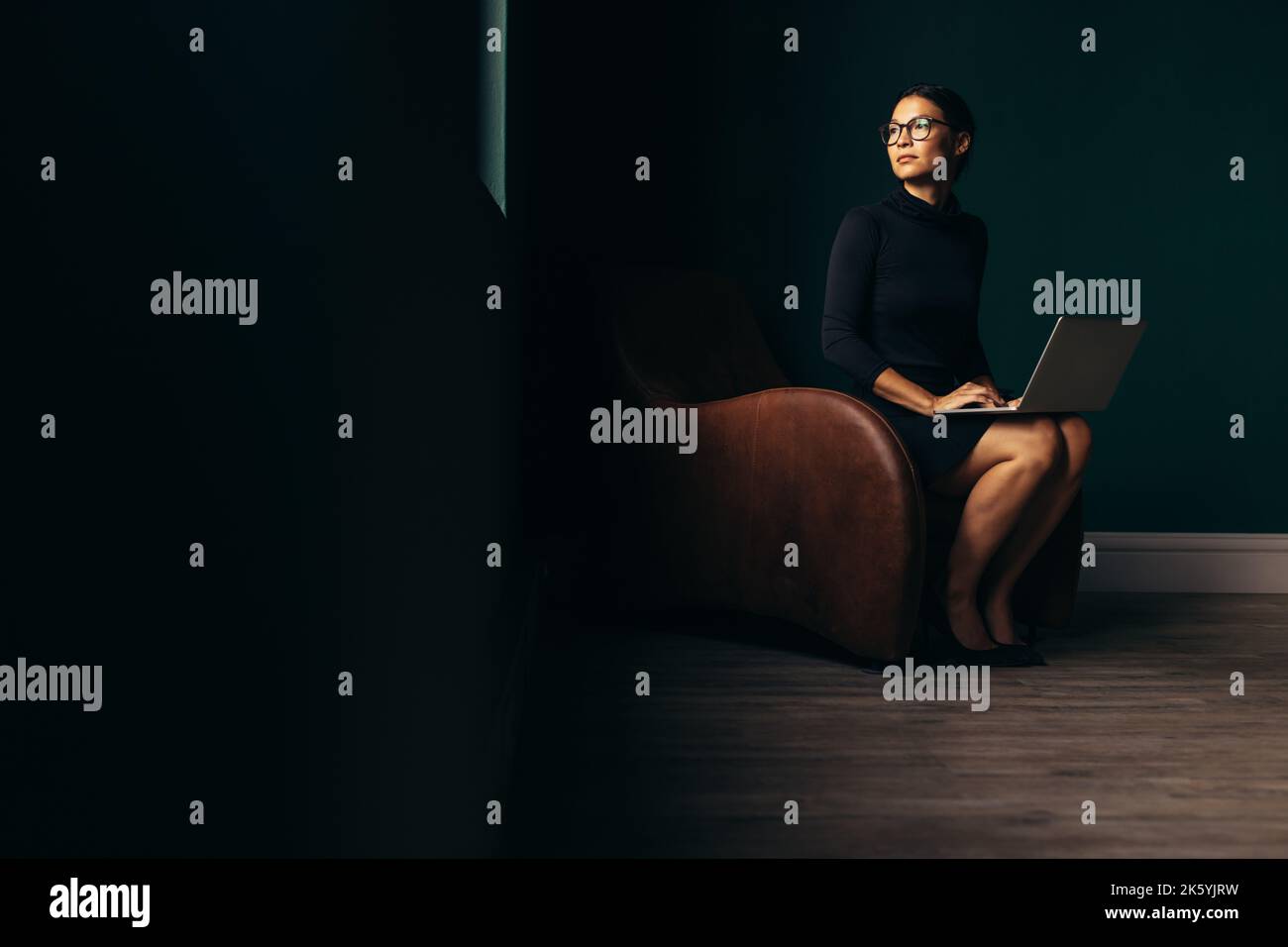 Donna premurosa seduta sulla poltrona con il computer portatile e guardando lontano. Giovane donna che lavora sul notebook e guarda fuori dalla finestra. Foto Stock