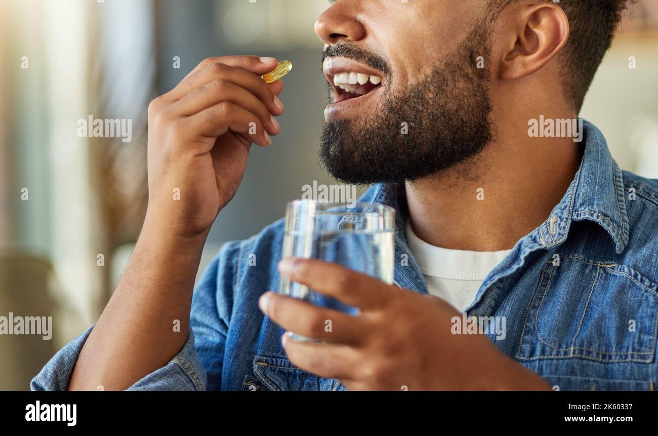 Bachelor bere farmaco con un bicchiere d'acqua. Uomo ispanico che beve una capsula di supplemento nel suo appartamento. Uomo che usa un bicchiere d'acqua per prendere Foto Stock