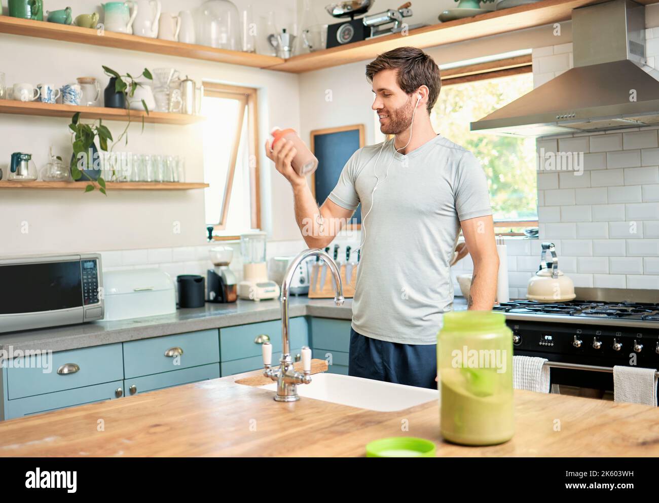 Un giovane uomo caucasico vestito bottiglia di proteine di siero di cioccolato agitare per l'energia per l'allenamento mentre si indossano gli auricolari in una cucina a. Foto Stock