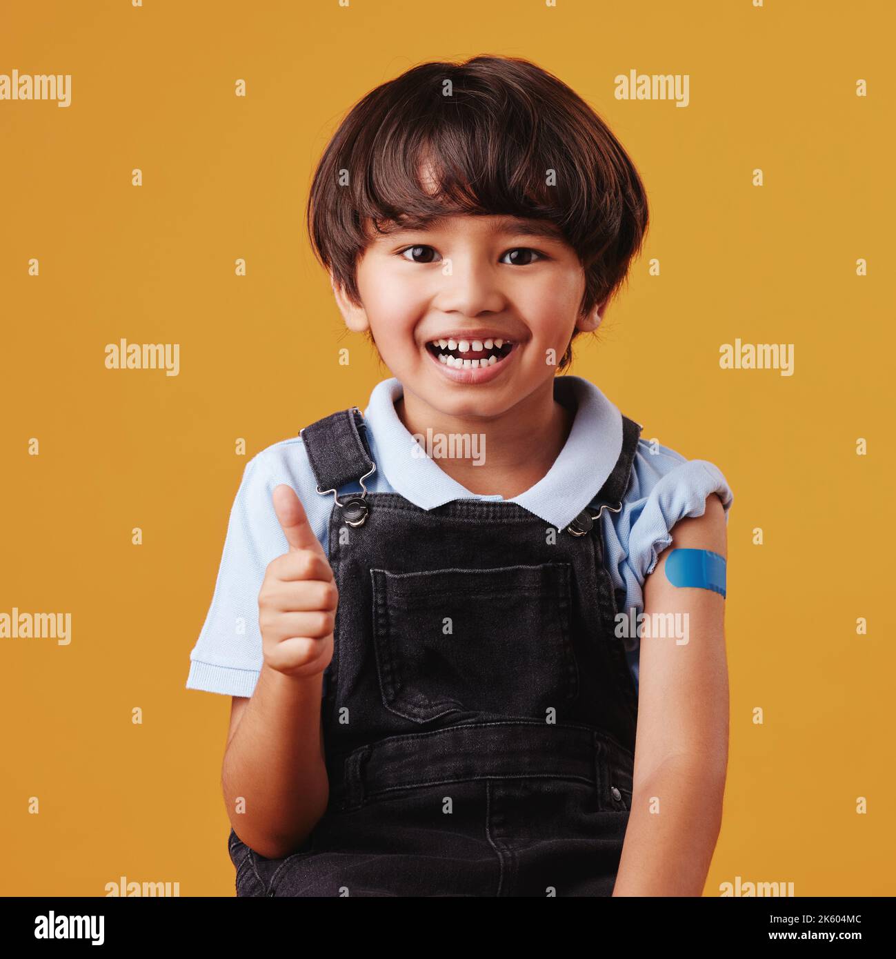 Ritratto di un sorridente felice bambino di razza mista che dà i pollici fino al vaccino mentre mostra il suo bandaid e posa contro un copyspace arancione Foto Stock