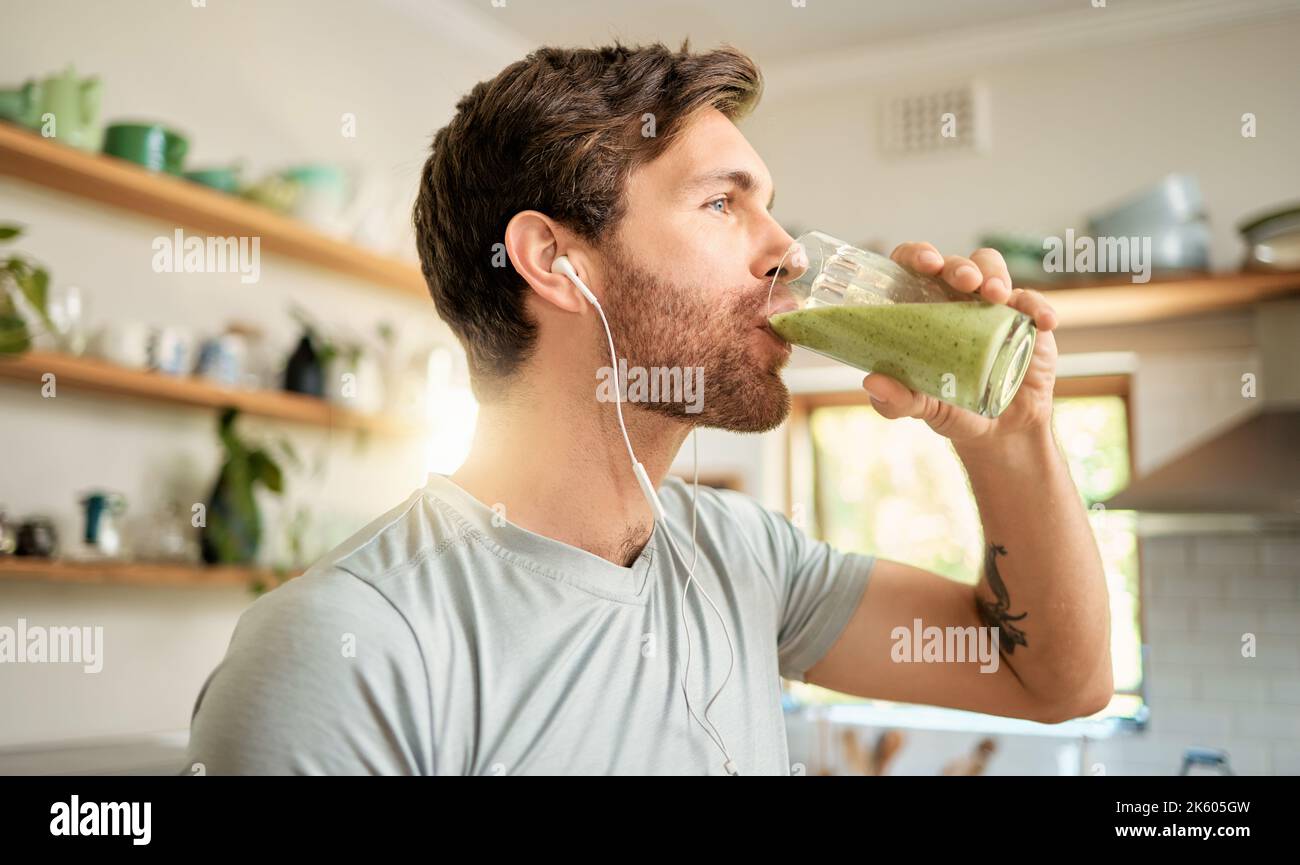 Un giovane uomo caucasico in forma bere un bicchiere di sano smoothie verde disintossicante mentre si indossano gli auricolari in una cucina a casa. Ragazzo che ha frutta fresca Foto Stock