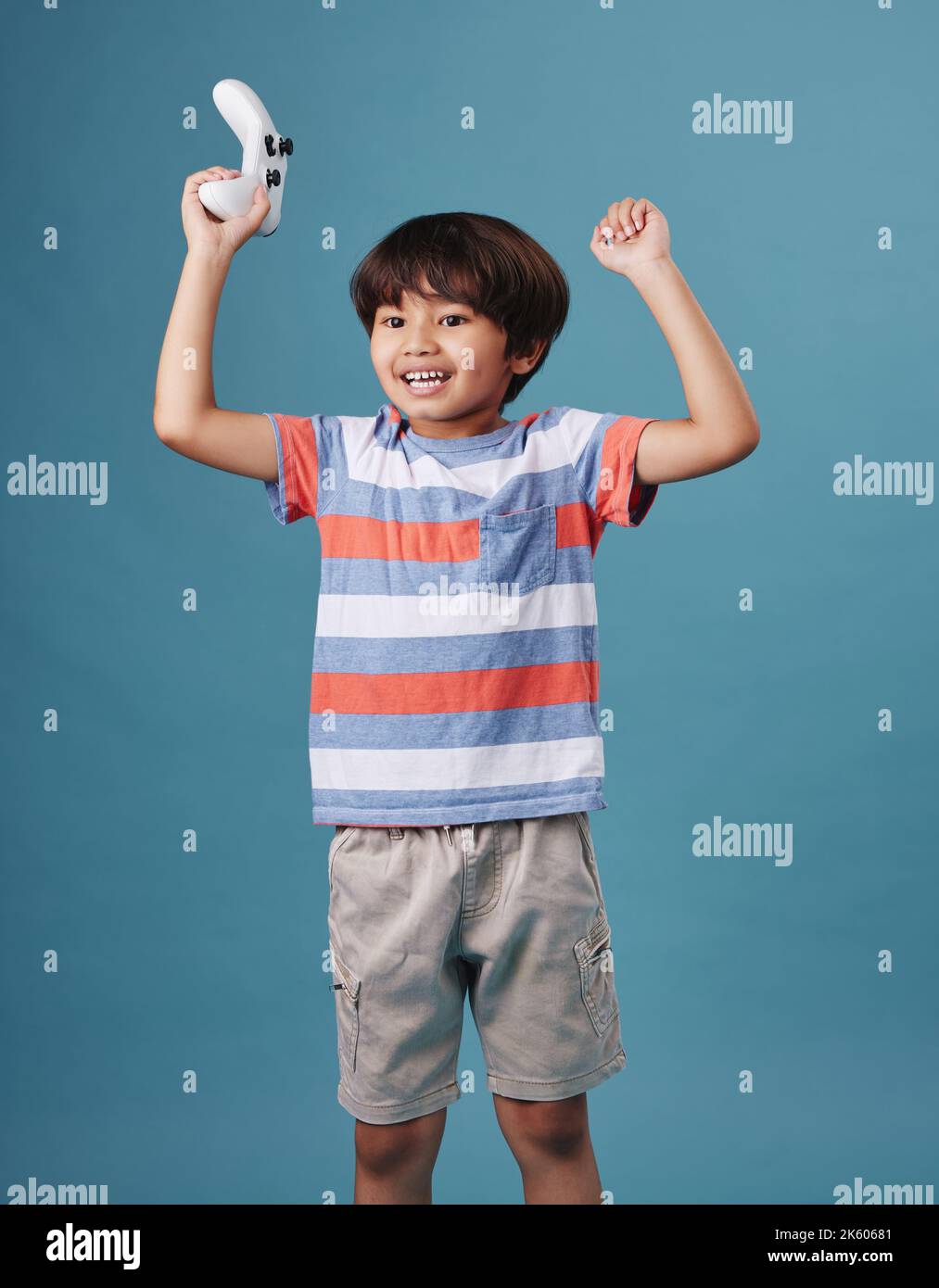 Giovane ragazzo misto da corsa in piedi e tenendo un controller console mentre si gioca un videogioco su sfondo blu. Bambino simpatico che celebra la vittoria a. Foto Stock