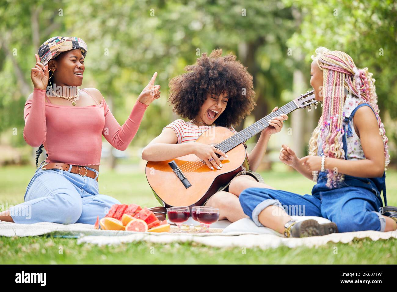 Donne nere, amici e pic-nic nel parco con chitarra suonando musica, cantando e trascorrere il tempo insieme. Fumetto, divertente e felice Signore con acustico Foto Stock