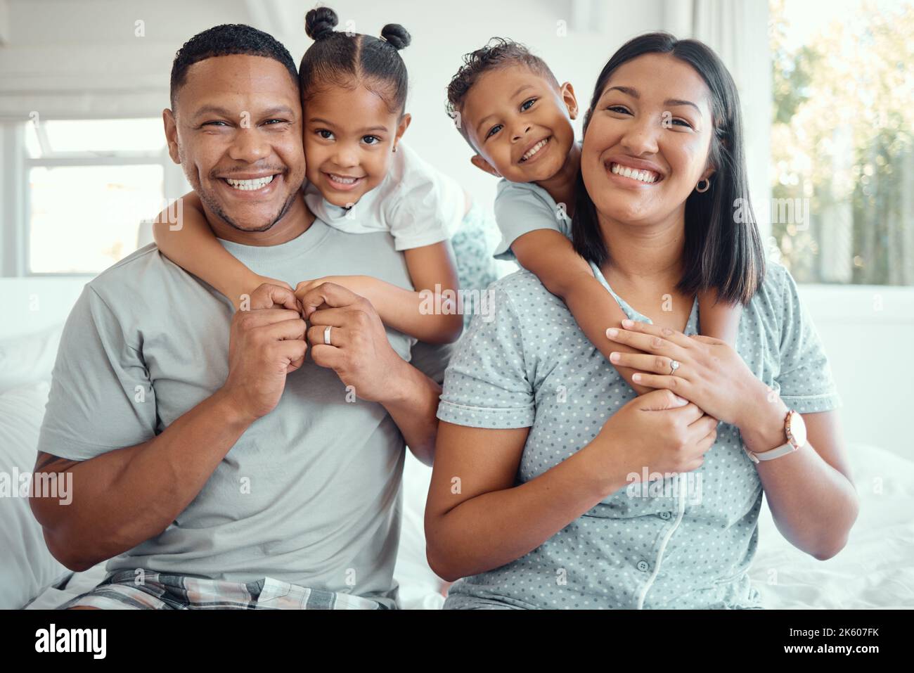 Ritratto di una giovane e felice famiglia mista gara con due bambini che indossano pigiami e seduti a casa insieme. Carino ragazza e ragazzo abbracciando loro Foto Stock