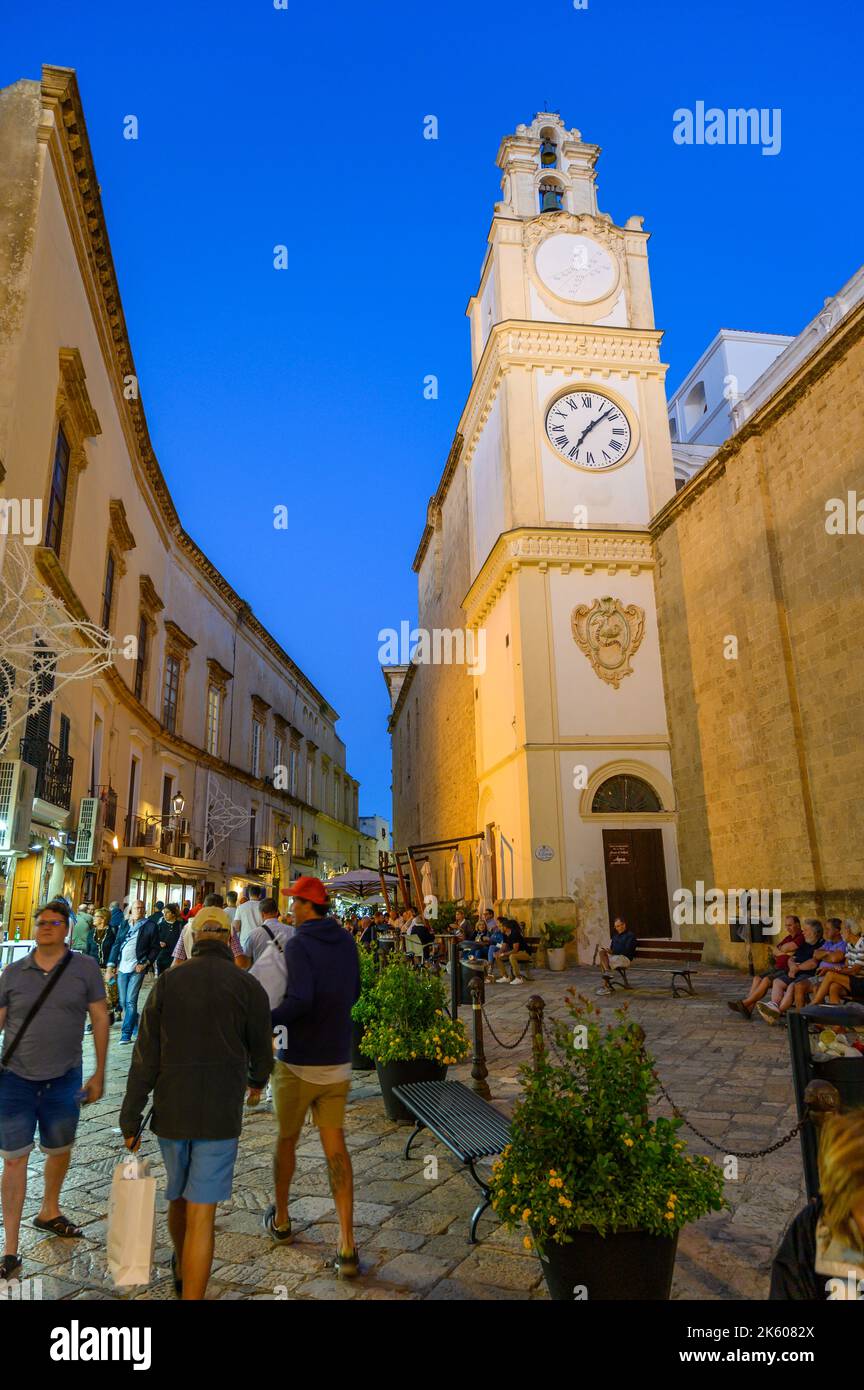 Turisti e locali passeggiando davanti alla torre dell'orologio della Cattedrale di Gallipoli su una strada acciottolata nel centro storico di Gallipoli in serata, Puglia, Italia. Foto Stock