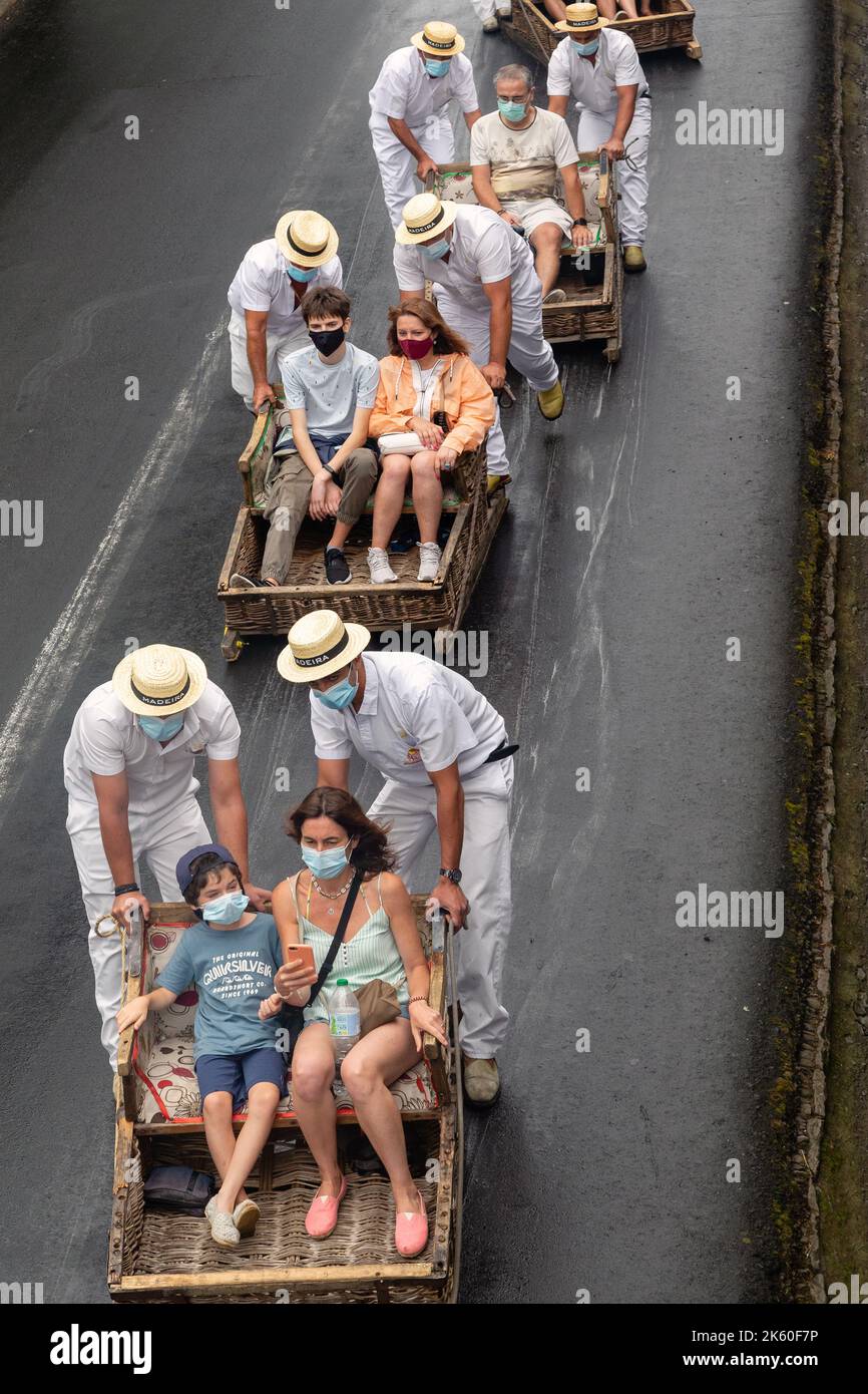FUNCHAL, PORTOGALLO - 24 AGOSTO 2021: Un gruppo di turisti non identificati rotolare giù per la strada in cesti di vimini (toboga) con l'aiuto dei conducenti in t Foto Stock