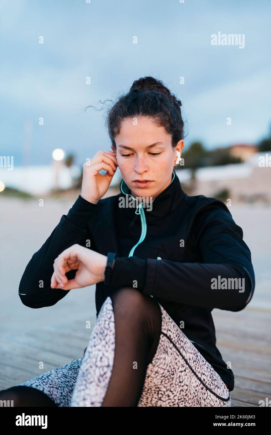 Giovane donna in abbigliamento sportivo con smartwatch e ascoltando musica con le cuffie prima di allenarsi in spiaggia Foto Stock