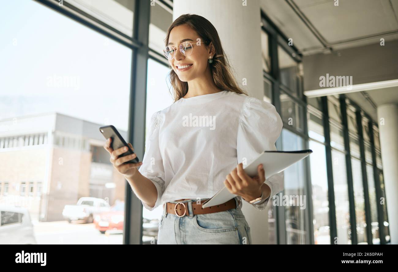 Felice donna d'affari che sta inviando messaggi sullo smartphone tenendo file e camminando in un ufficio moderno. Sorridere un'imprenditrice femminile utilizzando un'app mobile o navigando sui social network Foto Stock