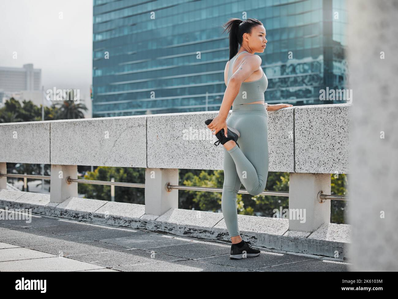 Una giovane donna in forma che indossa abiti da ginnastica e allunga le gambe mentre si trova in piedi su un ponte e guarda la vista della città. Atleta ispanico Foto Stock