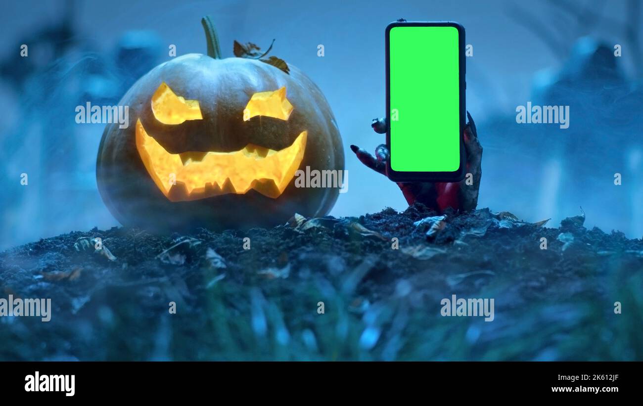 La mano di Zombie tiene lo smartphone con lo schermo verde fuori dalla tomba vicino alla zucca di Halloween incandescente nel cimitero. Evento di festa Halloween concetto. Foto Stock