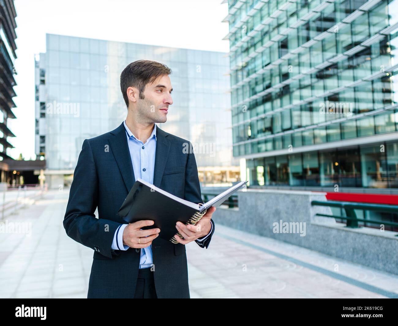Imprenditore maschio in elegante vestito che tiene il blocco note e guardando via sognatamente sulla strada soleggiato contro l'edificio contemporaneo Foto Stock