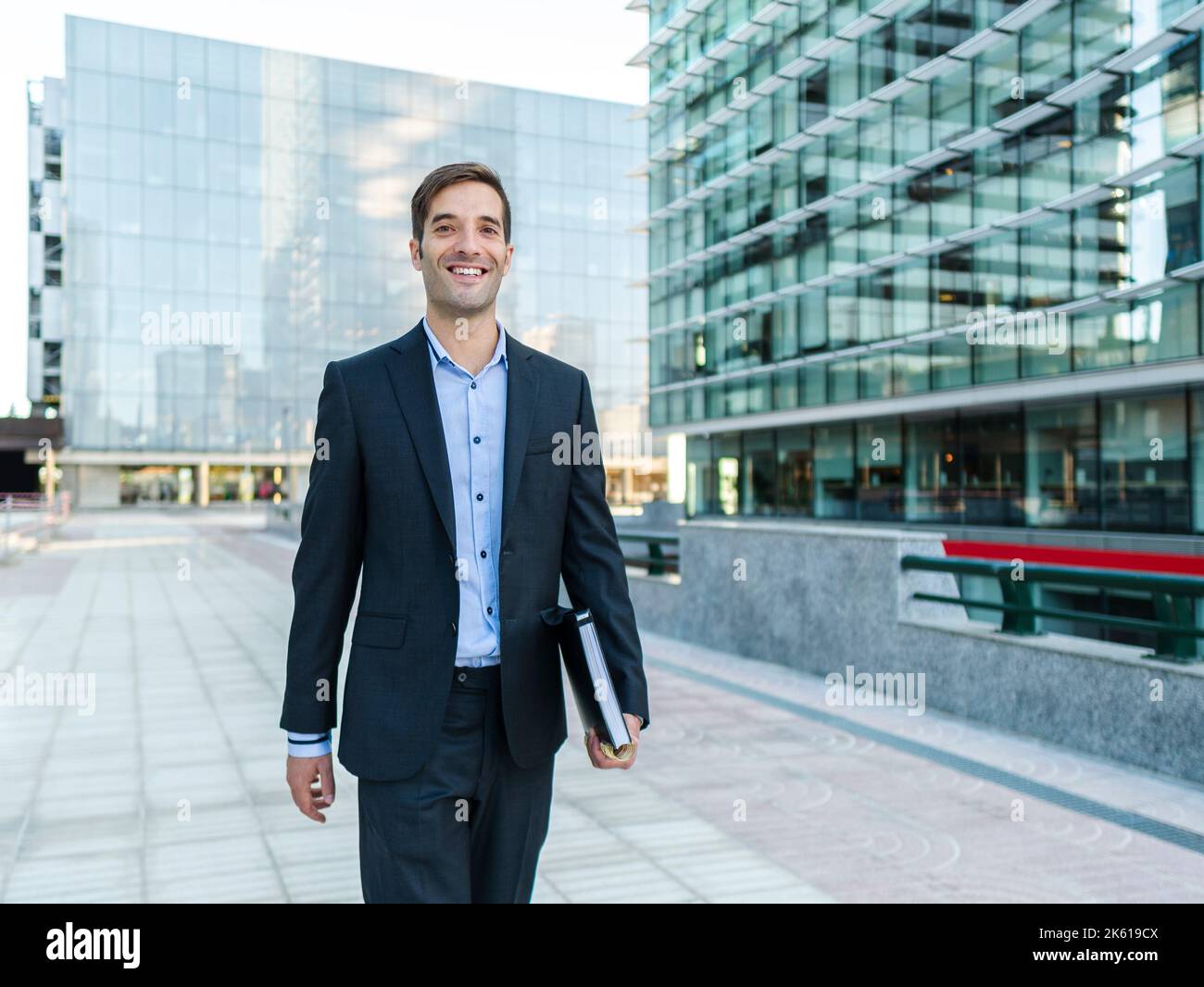 Imprenditore maschio in elegante vestito che tiene il blocco note e guardando la macchina fotografica sulla strada soleggiata contro l'edificio contemporaneo Foto Stock