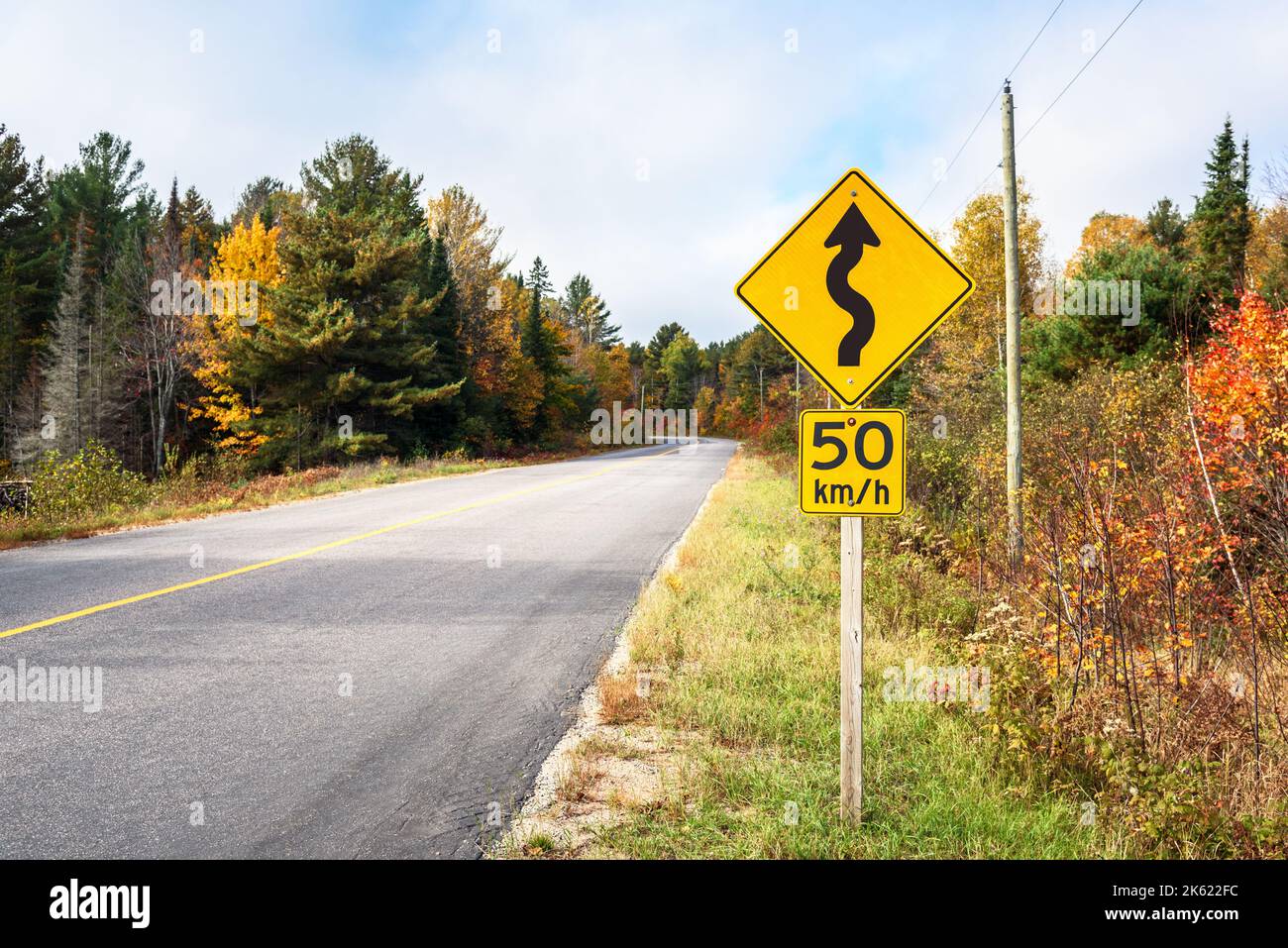 Cartello stradale di avvertimento lungo una strada forestale tortuosa in una giornata di sole autunnale. Splendidi colori autunnali. Foto Stock