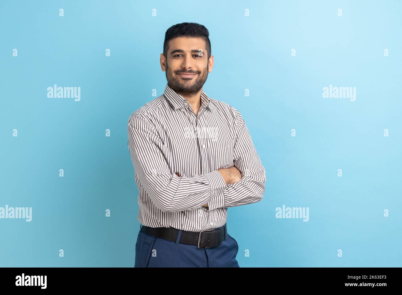 Giovane uomo d'affari felice e di successo che si posa con felice espressione fiduciosa, guardando la macchina fotografica, tenendo le braccia piegate, indossando una camicia a righe. Studio in interni isolato su sfondo blu. Foto Stock