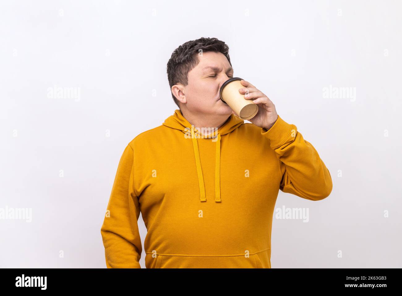 Ritratto di un bell'uomo di mezza età in piedi e bere un caffè take away in una tazza di carta, stile di vita fresco, con felpa con cappuccio in stile urbano. Studio in interni isolato su sfondo bianco. Foto Stock