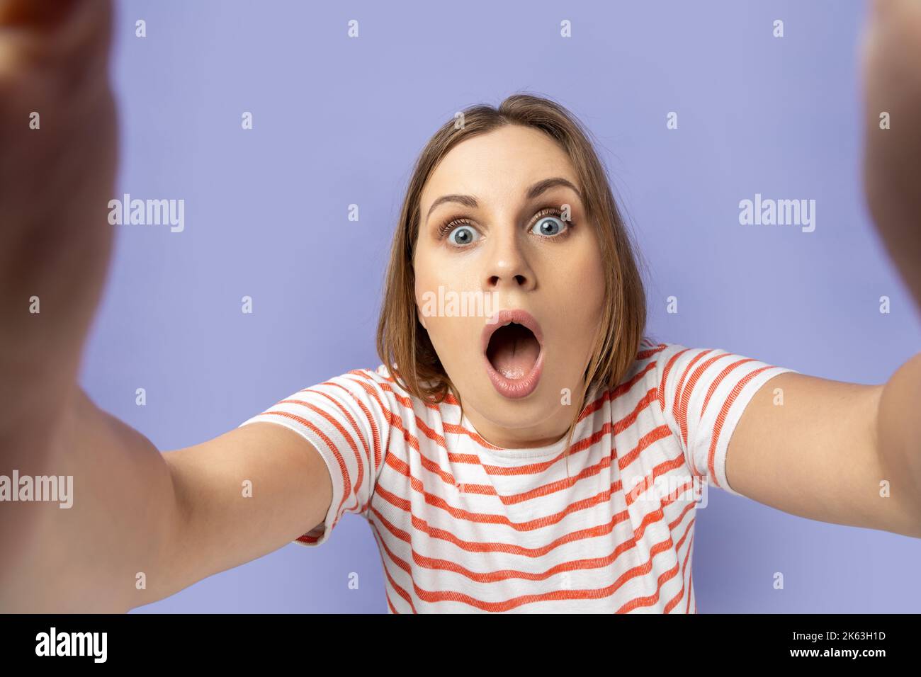 Ritratto di donna scioccata in T-shirt a righe scattando foto selfie, punto di vista, guardando la fotocamera con bocca aperta, facendo autoritratto anteriore. Studio al coperto isolato su sfondo viola. Foto Stock