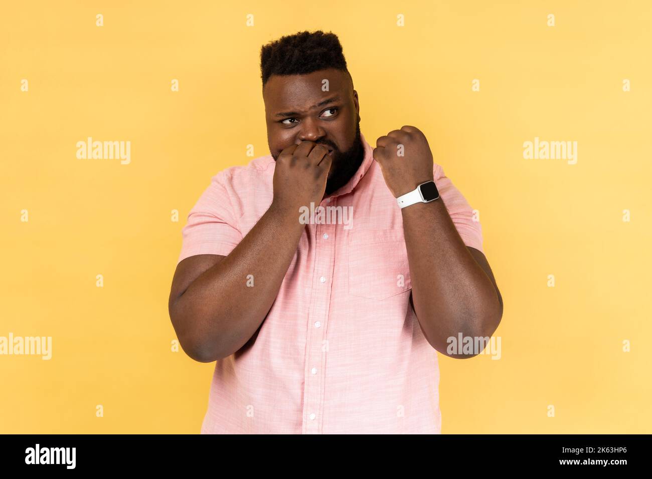 Ritratto di nervoso uomo bearded che indossa una camicia rosa mostrando il suo orologio intelligente e mordendo le unghie delle dita, non ha abbastanza tempo. Studio al coperto isolato su sfondo giallo. Foto Stock