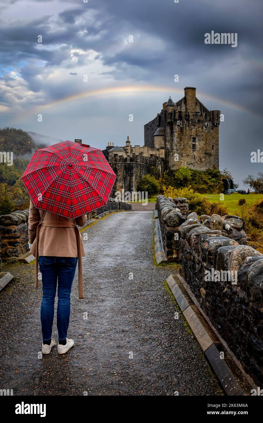 Una donna turistica con un ombrello scozzese cammina verso il castello di Eilean Donan Foto Stock