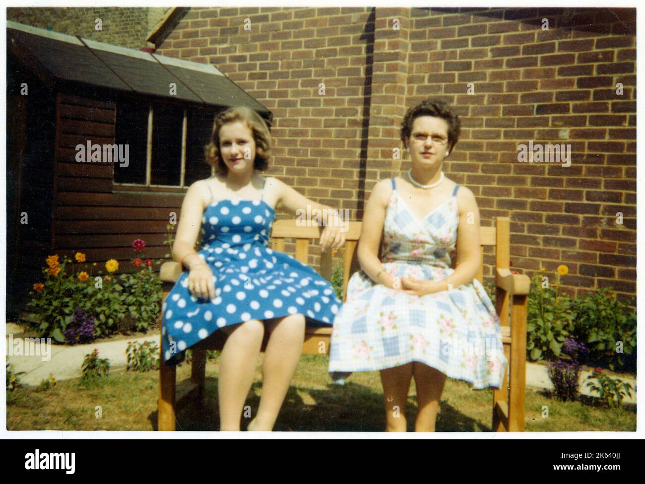 Una madre e un duaghter nei loro migliori camici estivi che si affacciano su una panchina in un giardino suburbano molto pulito e curato retro - Agosto, 1959. Foto Stock
