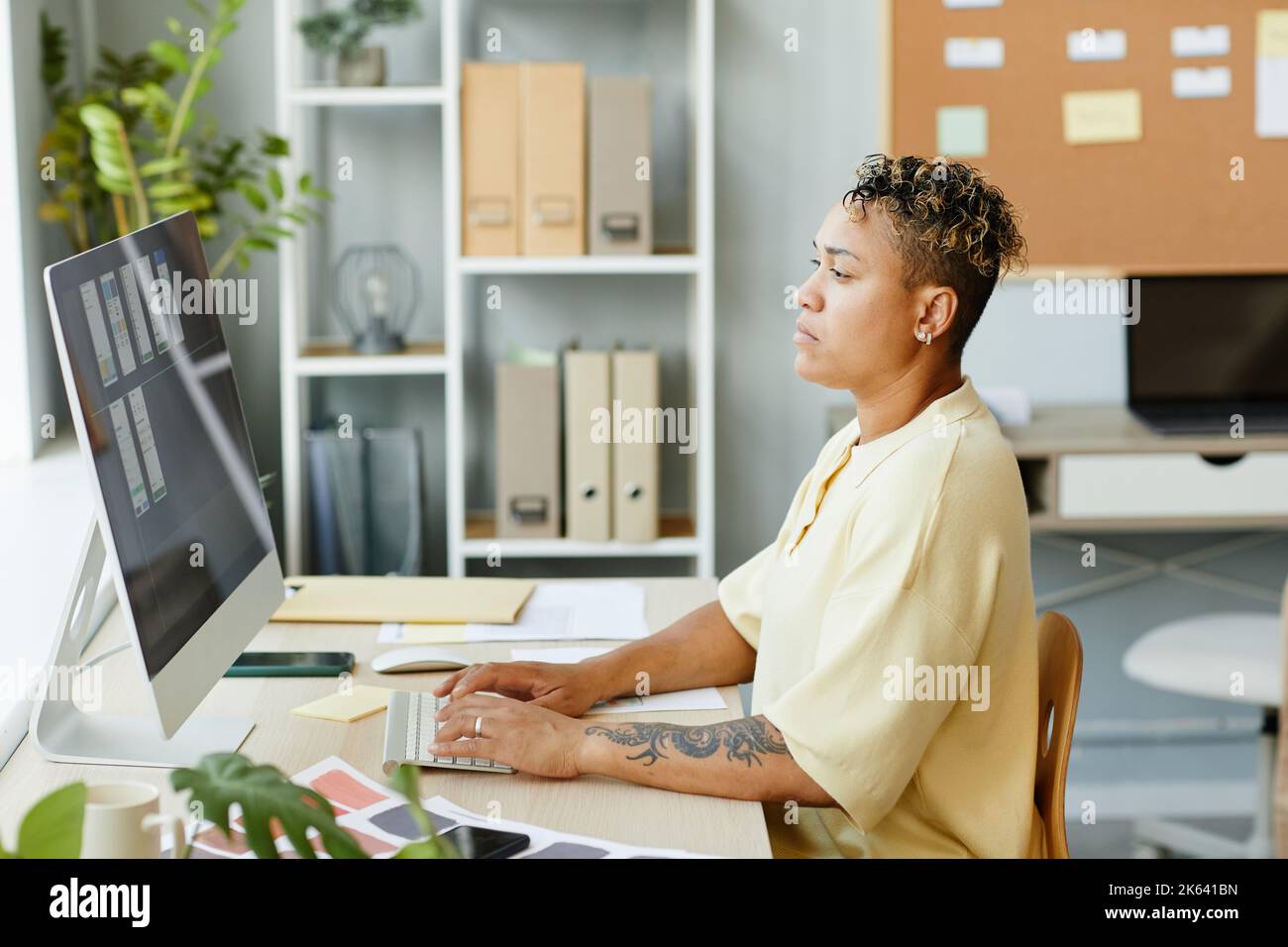 Vista laterale ritratto di una donna nera tatuata che utilizza il computer in ufficio mentre progetta l'interfaccia app mobile Foto Stock