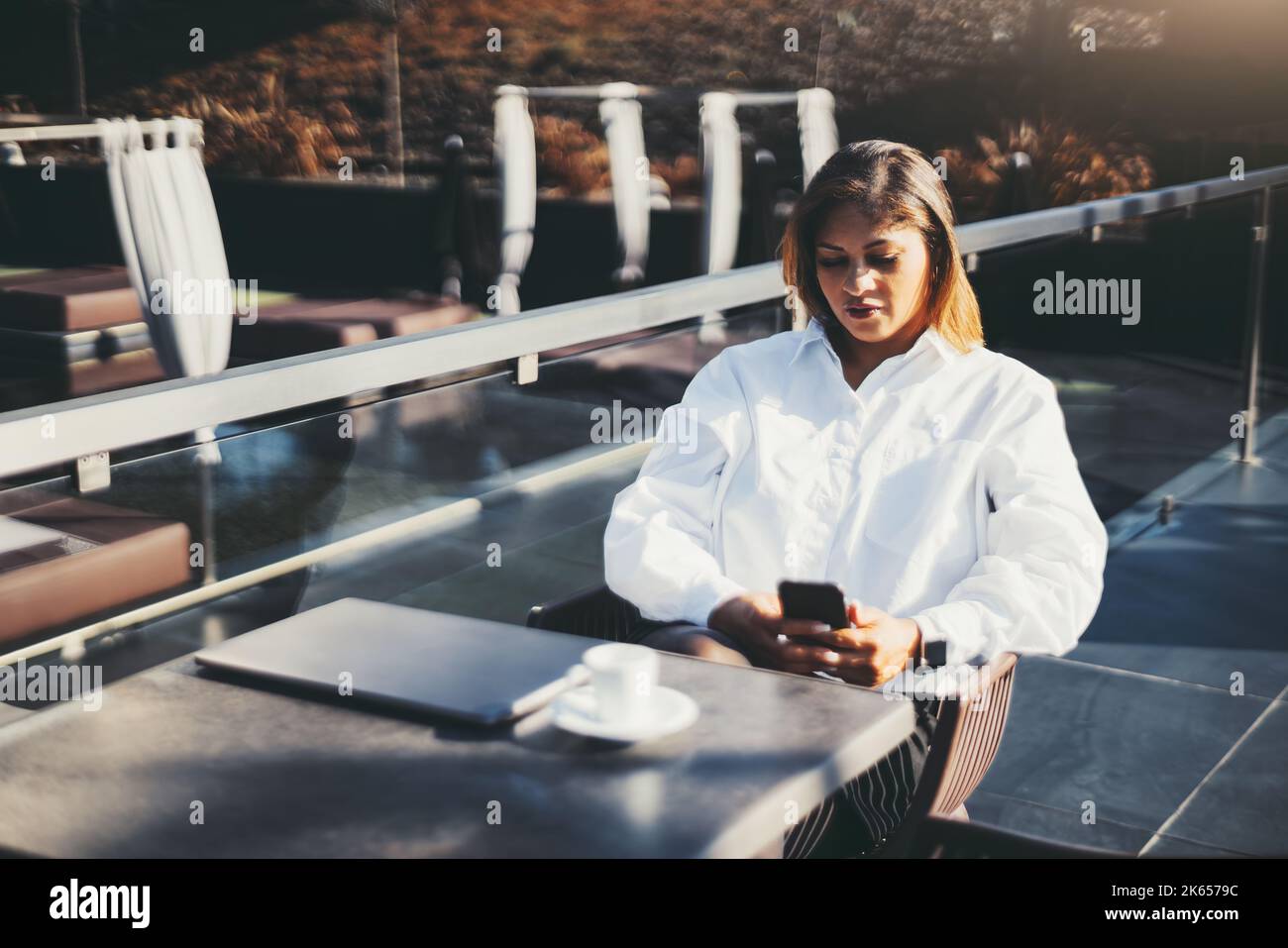 Vista di una tranquilla e affascinante donna d'affari ispanica seduta al tavolo di un ristorante all'aperto da solo dopo il lavoro e la lettura dei messaggi sul suo smartphone; Foto Stock