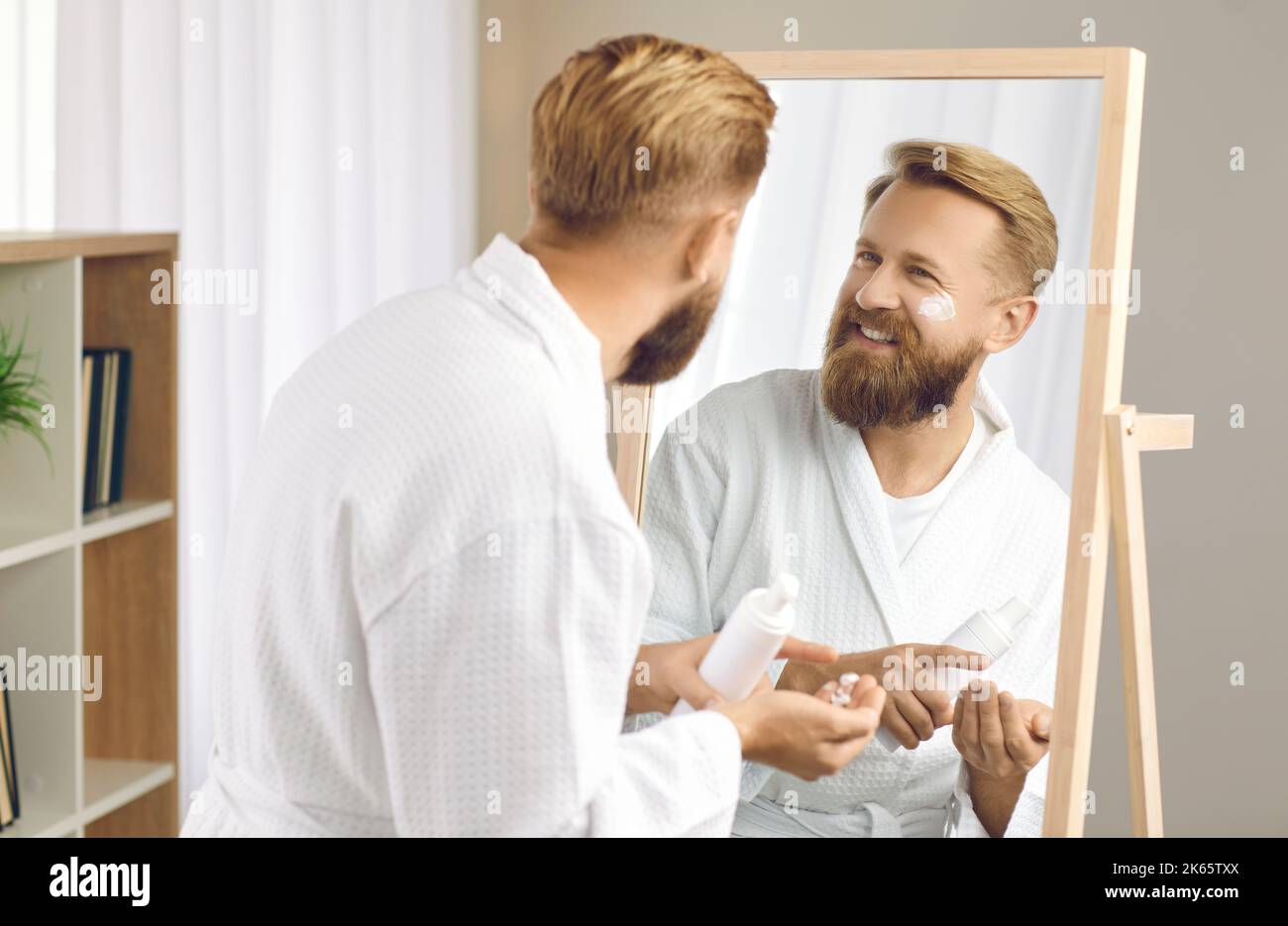 Uomo felice guardando il suo volto nello specchio mentre fa la sua routine di cura della pelle del mattino Foto Stock