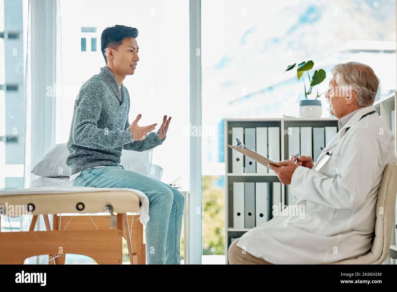 Un giovane asiatico che parla mentre si consulta con un medico caucasico senior mentre si siede su un letto in clinica. Paziente di razza mista che spiega come lui Foto Stock