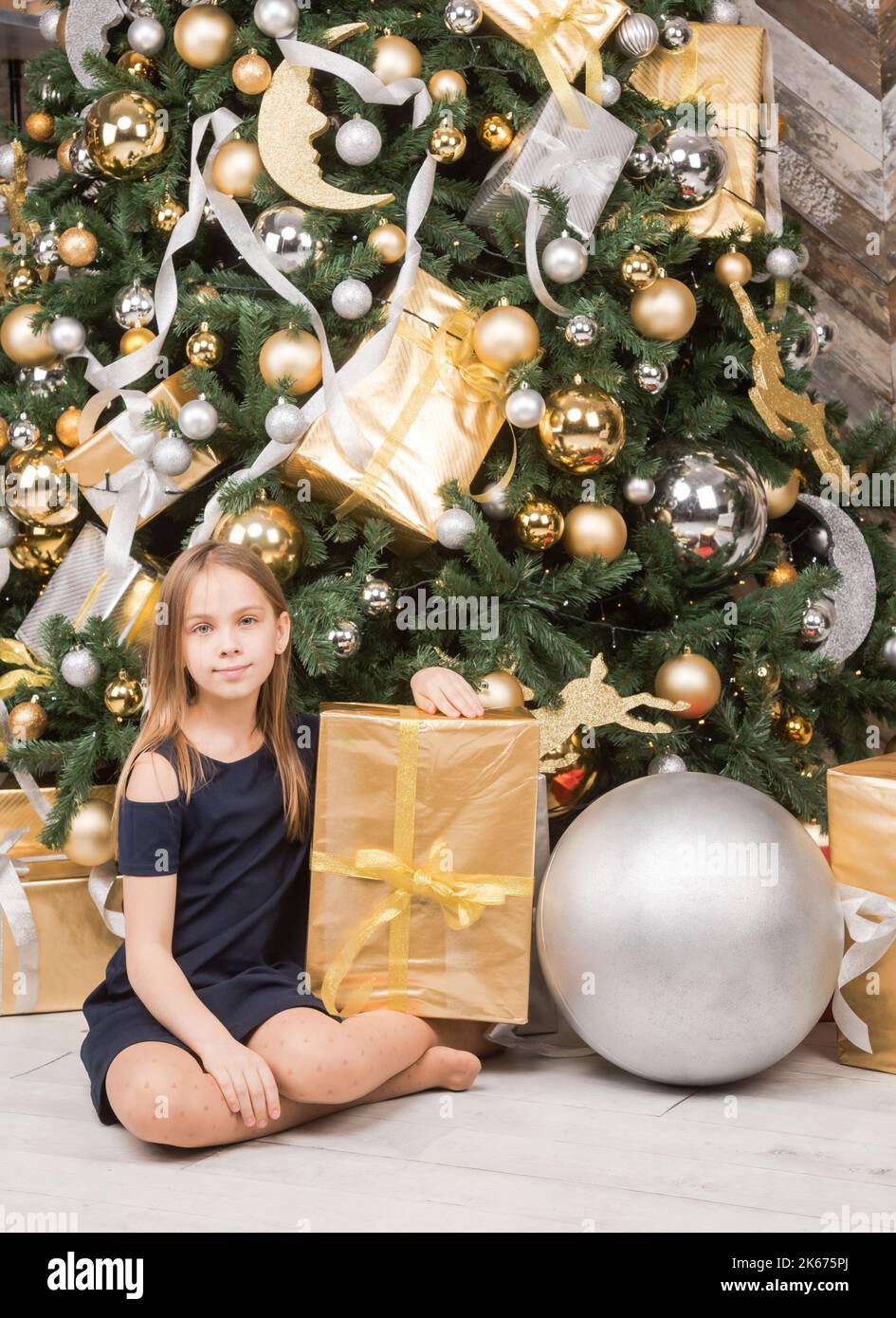 Ritratto di ragazza preteen indossa vestito blu seduto tra i regali impacchettati nei pressi di albero di Natale decorato felice di avere un sacco di regali sul Boxing Day Foto Stock
