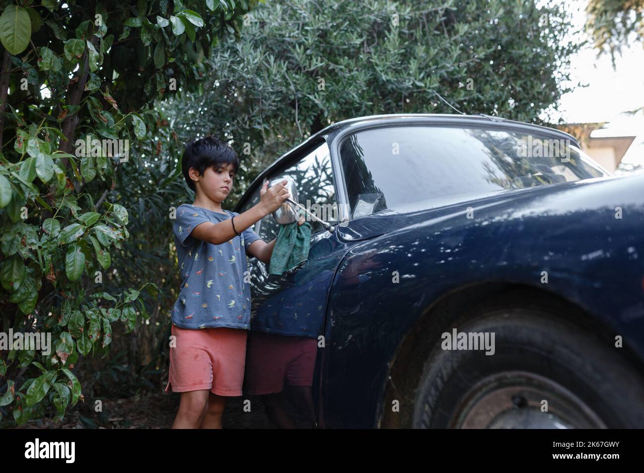 Ragazza di 8 anni con capelli corti e scuri che lavano lo specchio laterale di un'auto vintage blu Foto Stock