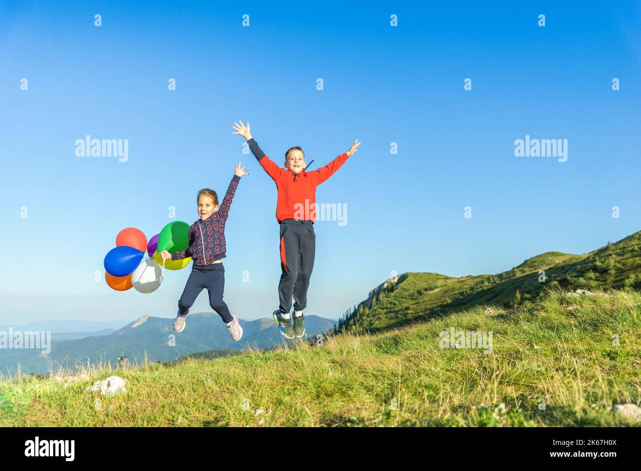 I bambini saltano con i palloncini in mano che si sballano nel vento. Vette di montagna e cielo blu sullo sfondo. Gioco, libertà e felicità dei bambini Foto Stock