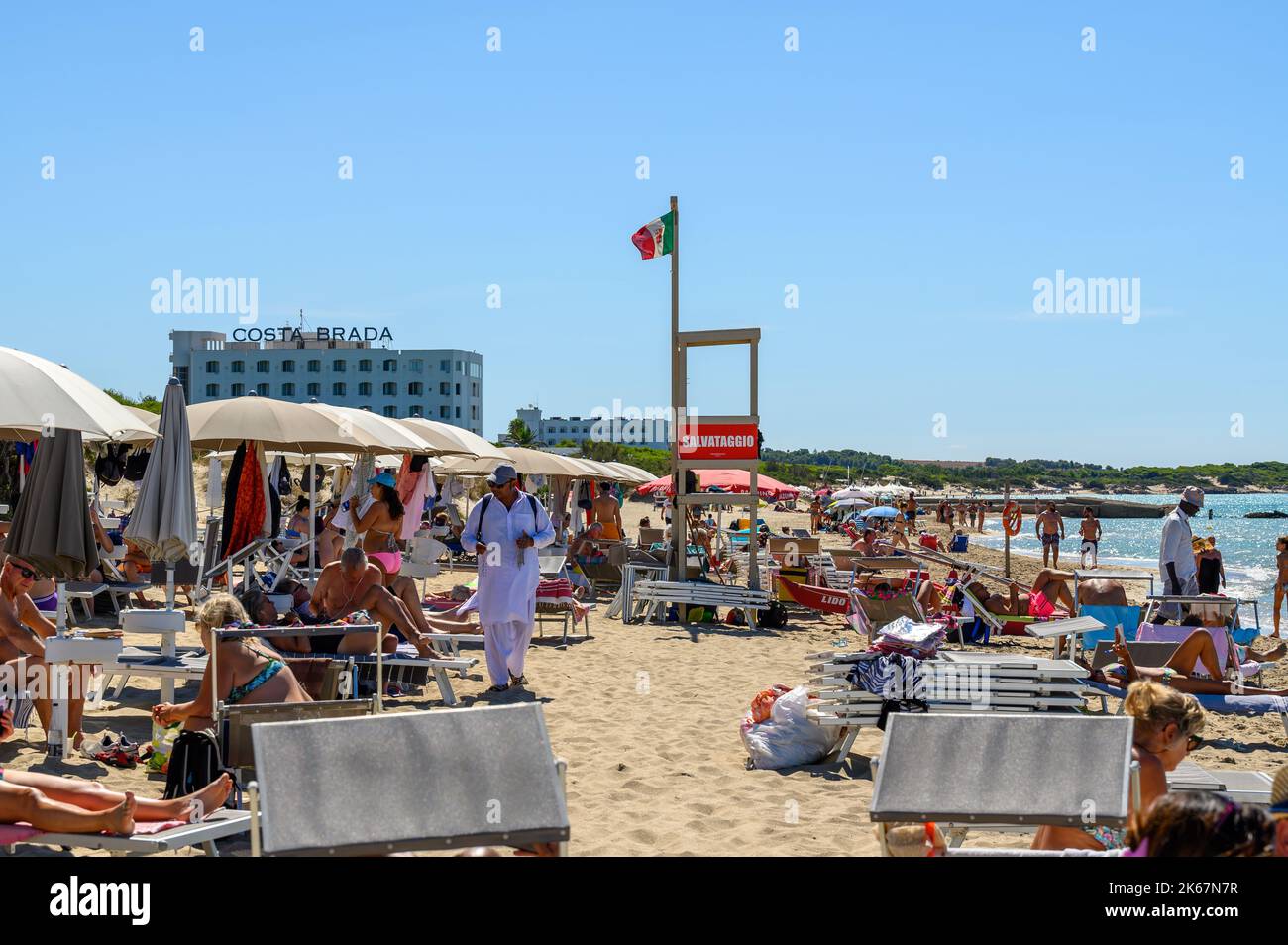 Un venditore di spiagge cammina tra i bagnanti e i beachgoer sulla spiaggia di Sottovento cercando di vendere collane. Vicino a Gallipoli in Puglia, Italia. Foto Stock