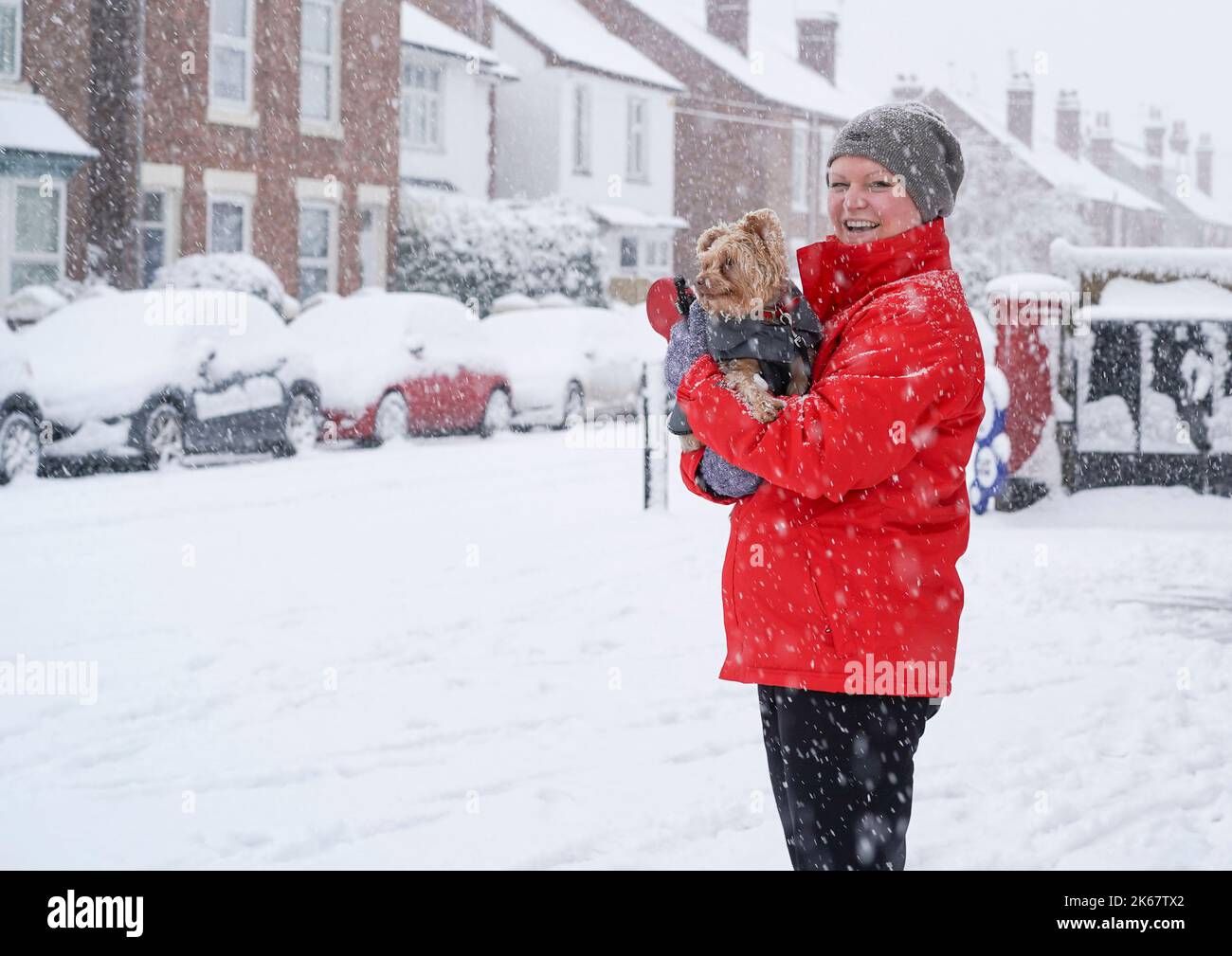 Donna sorridente nella neve pesante, indossando un cappotto rosso, tenendo il cane tra le braccia sul lato di una strada mentre nevica. Foto Stock