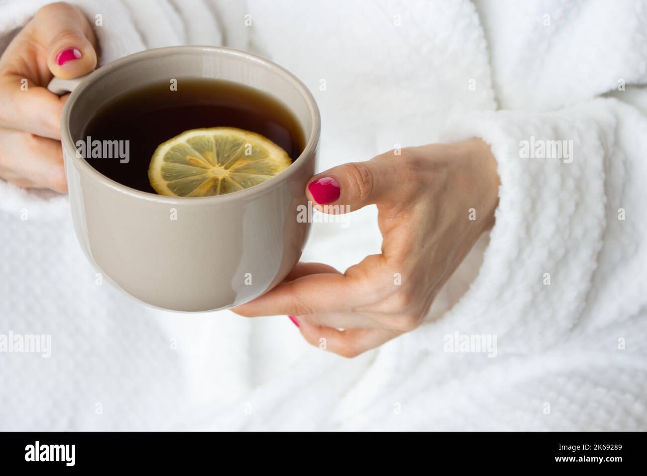 Donna che tiene una tazza di tè con limone. Ragazza in accappatoio bianco con tazza di tè marrone. Concetto di comfort domestico. Concetto di bevanda mattutina. Relax e comfort. Foto Stock