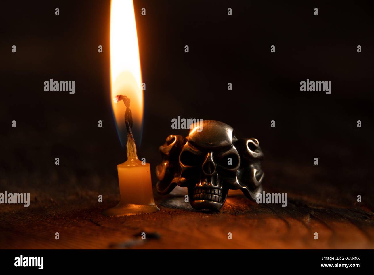 Anello d'argento a forma di cranio accanto ad una fiamma di candela su un tavolo di legno al buio, il cranio è d'argento Foto Stock